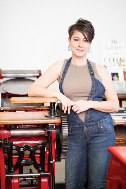 Julie Wall stands in blue jean overalls next to her red printing press.